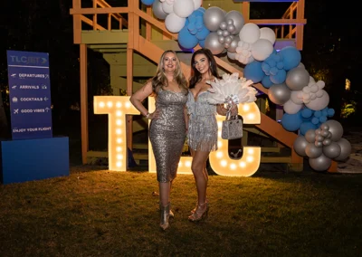 Two women in sparkling silver dresses in front of lifeguard stand adorned with balloons.