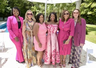 Six women in colorful dresses smiling outdoors near a pool and palm trees.